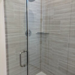 Glass shower with gray tile walls, a metal showerhead, a handle, and a small corner shelf.