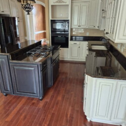 A spacious kitchen with white cabinets, black countertops, an island stove, double oven, and wood flooring.