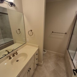 Bathroom with a single sink vanity, mirror, towel rings, bathtub with glass panel, and beige tiled floor.