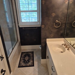 Bathroom with a white sink vanity, large mirror, tiled tub, small rug, and window with white shutters.