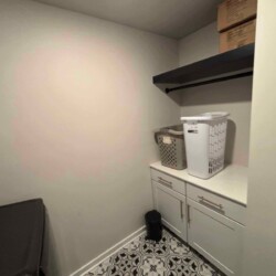 Small laundry room with two baskets on a counter, a shelf with boxes above, and patterned tile flooring.