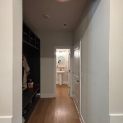 Hallway with wood floor, shelves on the left, and a bathroom with a pedestal sink and round mirror at the end.