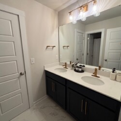 Modern bathroom with double sinks, black cabinets, gold fixtures, large mirror, and white tiled floor.