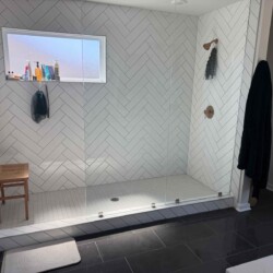 Large walk-in shower with white herringbone tile, glass door, wooden bench, toiletries, window, and two wall hooks.