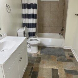 Bathroom with a white sink vanity, toilet, striped shower curtain, tiled tub, and multicolored tile floor.
